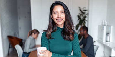 Brown hair women smiling, with other employees sitting at the table an office in the background