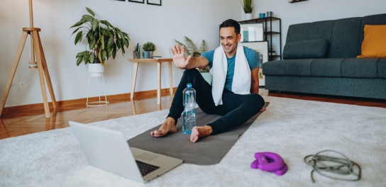 Men sitting on the ground and doing a home workout. Lapop is in front of him and he is sitting on a sports mat