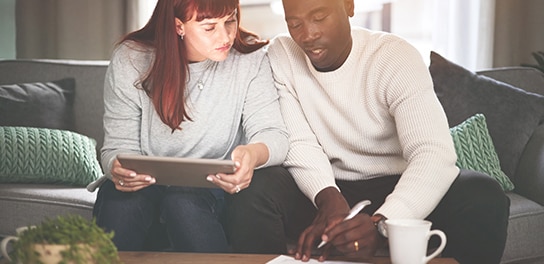 two people sitting on the coach. One person is writing soemthing downd and the other person is holding an iPad in the hand