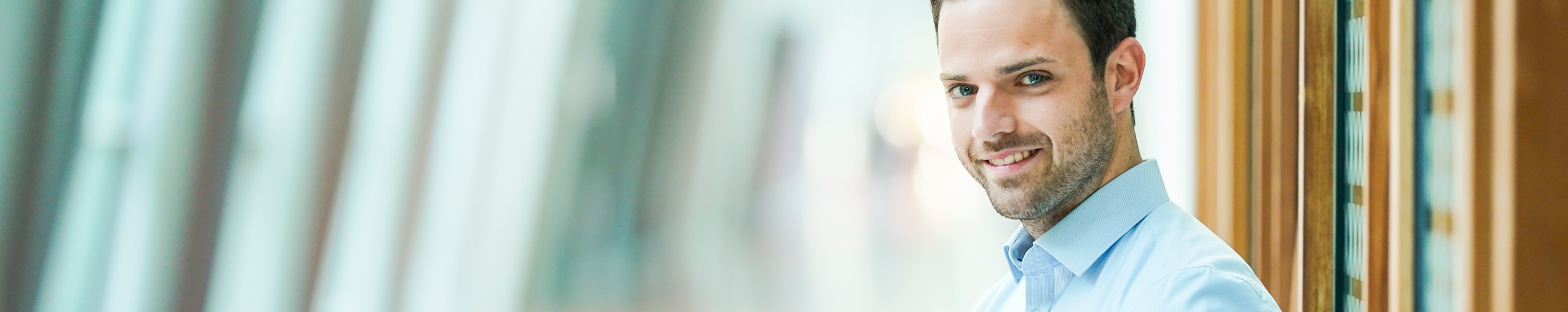 Ein Mann mit blauem Hemd lächelt und steht im Flur eines Bürogebäudes