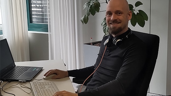 Mann sitzt an seinem Schreibtisch mit Kopfhörern und Laptop und einer Pflanze im Hintergrund