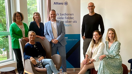 Ein Team der Allianz steht und sitzt in einem Büroraum und lächelt