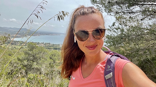 Frau mit Sonnenbrille, Sportkleidung und Kopfhörern beim Wandern mit Meer und Landschaft im Hintergrund