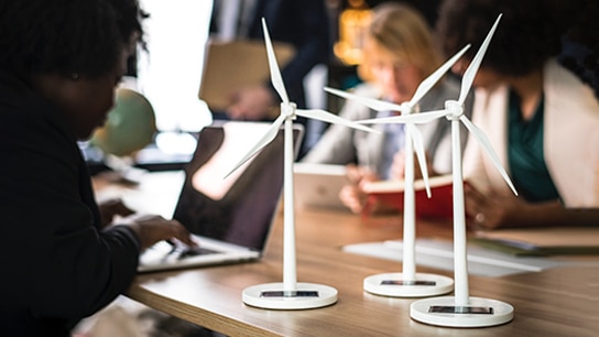 Close-up of miniature wind turbines on an office table, with colleagues working on laptops in the background.