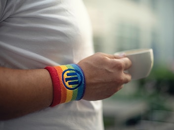A person's hand holding a cup, wears a rainbow coloured wrist band with Allian zlogo imprinted at the centre