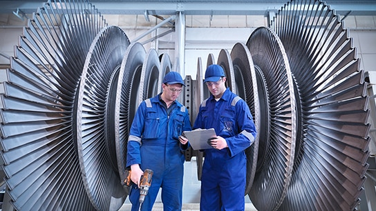 Two engineers are standing next to large machines, reviewing reports in one of their hands.