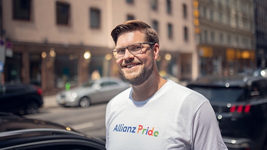 Mann steht auf einer Straße und lächelt und hat ein Allianz Pride Tshirt an