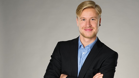 Mann hat einen Anzug an und steht vor einer grauen Wand
