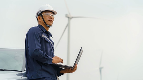 An engineer looking at the sky while using a laptop, with wind turbines in the background.