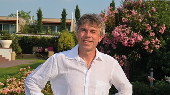 Mann in weißem Hemd steht vor einem Haus und einem Busch mit Blumen