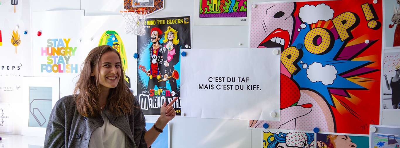 Une jeune femme regarde la caméra en souriant et montre du doigt un slogan accroché au mur