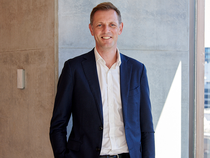 A man in a suit stands confidently in front of a large window, showcasing a professional demeanor and urban backdrop