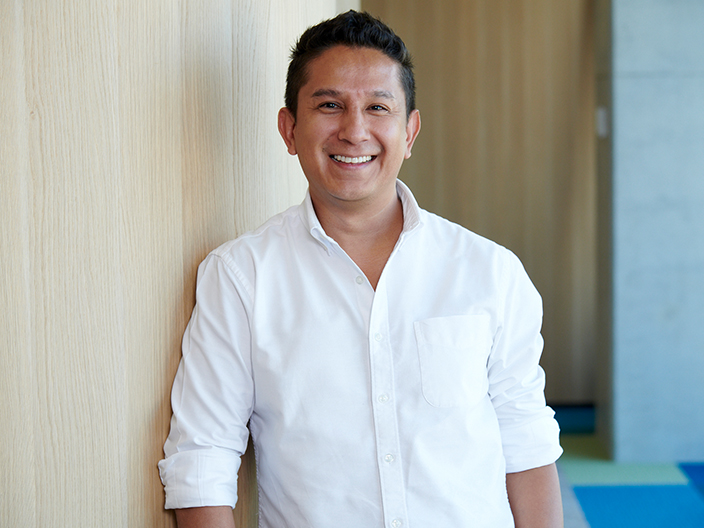 A smiling man in a white shirt stands confidently in front of a rustic wooden wall, exuding warmth and approachability