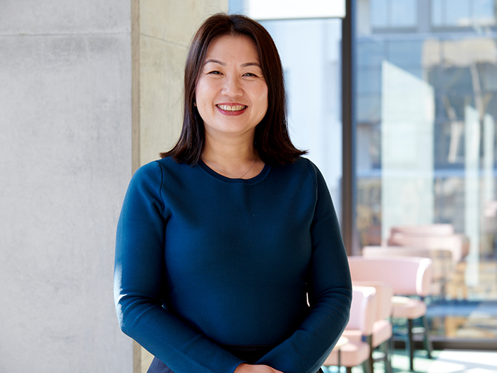 A woman in a blue shirt stands near a window, looking out with a contemplative expression