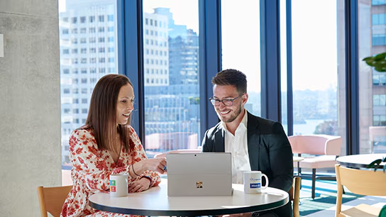 Two individuals seated at a table, engaged with a laptop, likely working together on a discussion or sharing ideas. 