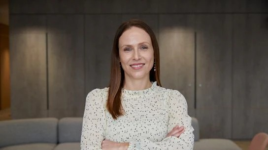 A woman with long hair wearing a white shirt, standing confidently against a neutral background 
