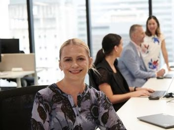 Employee smiles confidently, representing customer service, as team members work on laptops in the background, highlighting a professional and engaged workplace