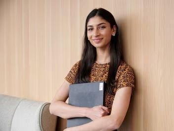 Young graduate with laptop leans on office wall, smiling, reflecting enthusiasm for her new professional journey