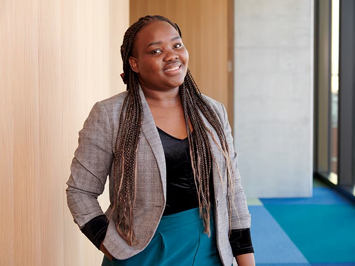 A woman with braids stands confidently in front of a textured wall