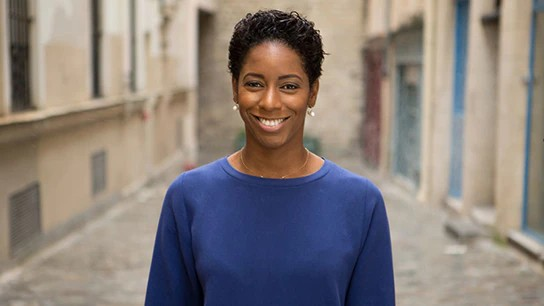 Malia wearing a blue shirt poses in an alley, with brick walls framing her against a backdrop of urban life