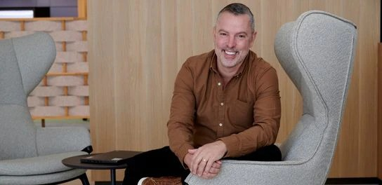 A man sits on a couch, smiling warmly and appearing relaxed