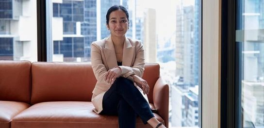 A woman seated on a couch, gazing out of a window, with natural light illuminating the room