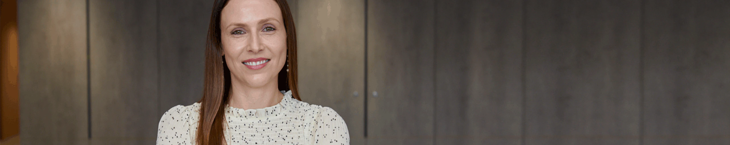 A woman with long hair wearing a white shirt, standing confidently against a neutral background