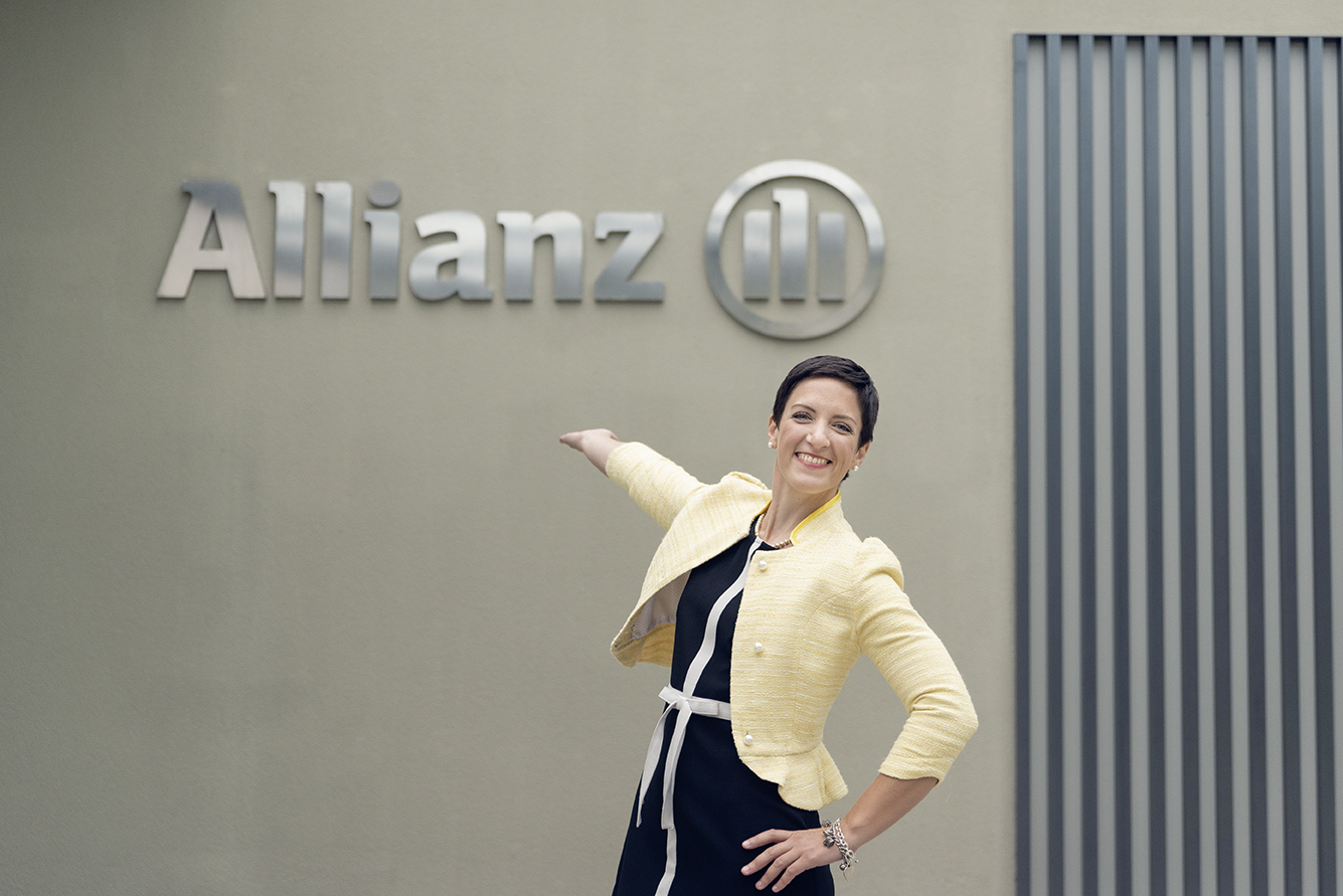 Allianz logo on a building wall with a person posing in front, wearing a light jacket and dark outfit.