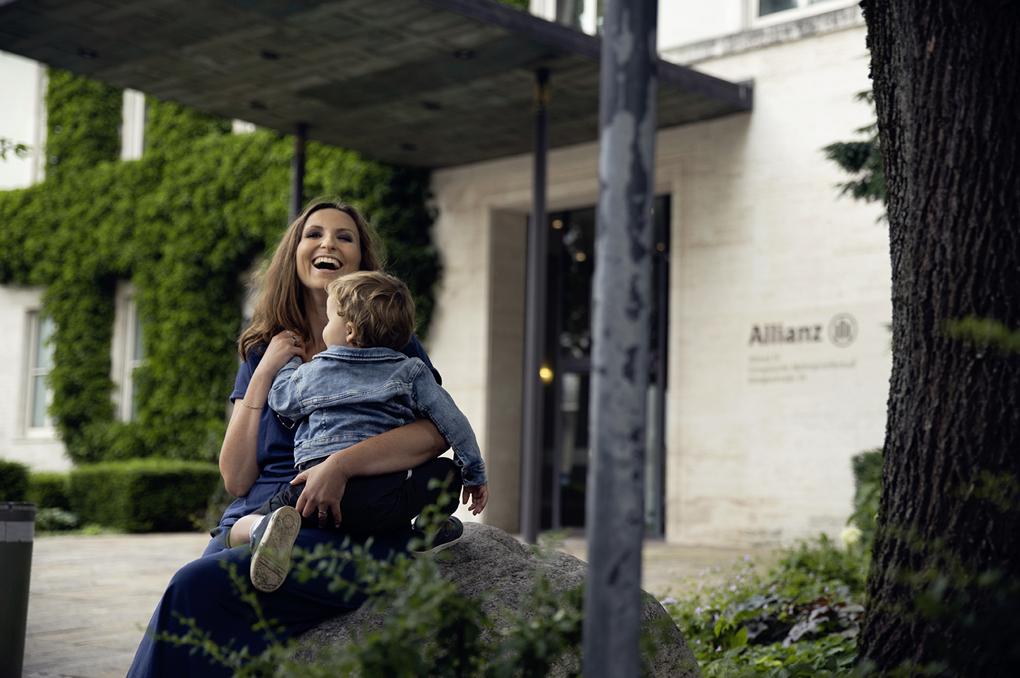 Person sitting outdoors on a stone bench surrounded by greenery, holding a child, with Allianz signage on the building in the background.