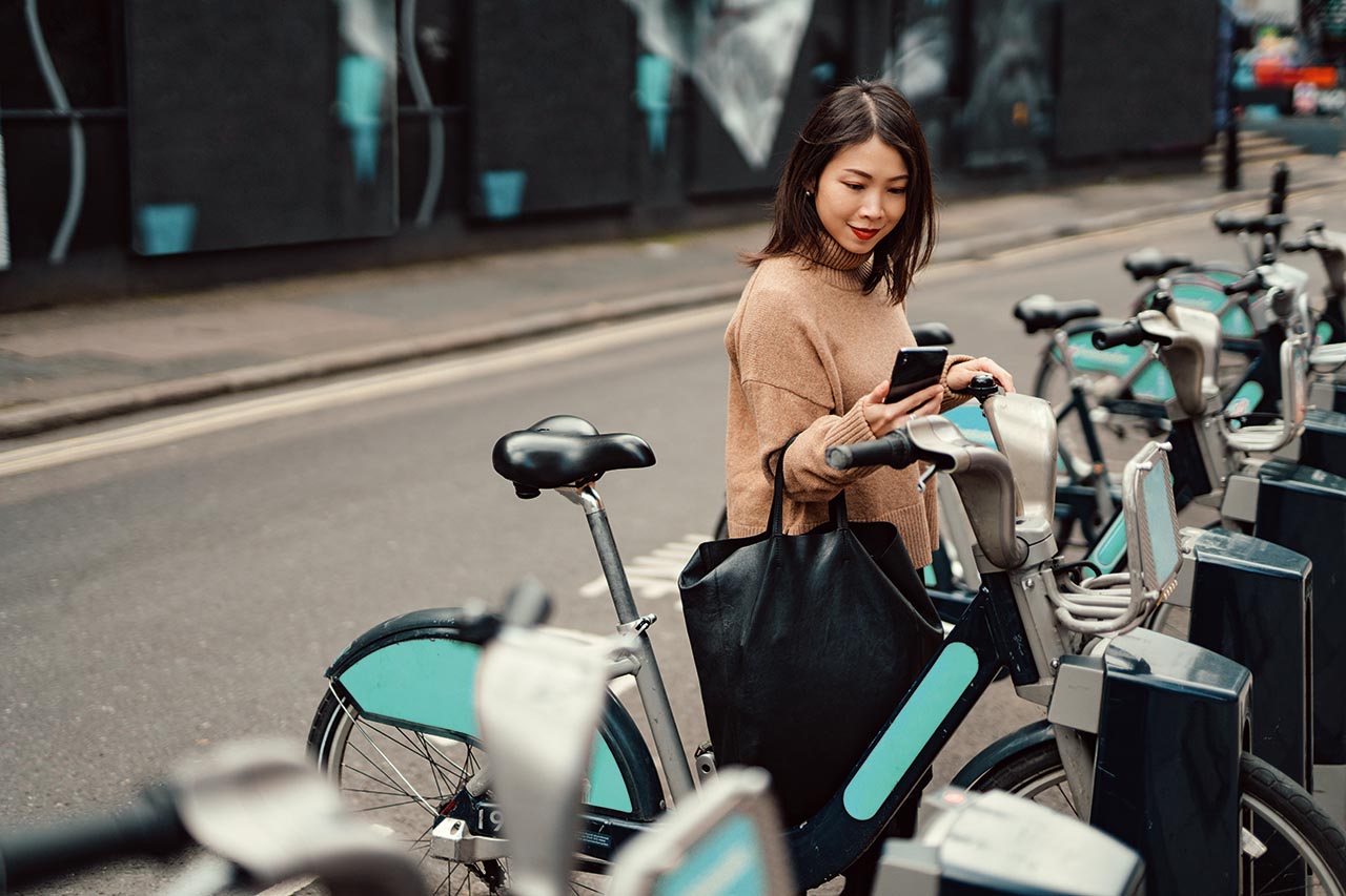 Young woman checking her phone while unlocking her bicycle at a public bike rack