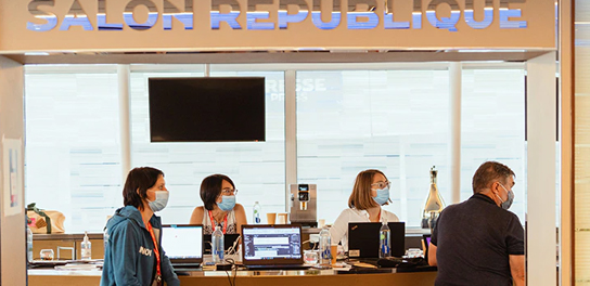 Des gens portant des masques, debout devant des ordinateurs portables dans un salon République, tous détournant le regard de la caméra.