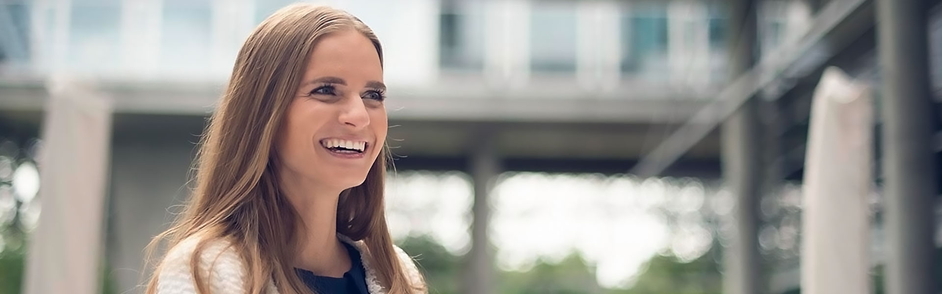 Person in a white textured jacket standing outdoors near a modern glass building with greenery in the background.