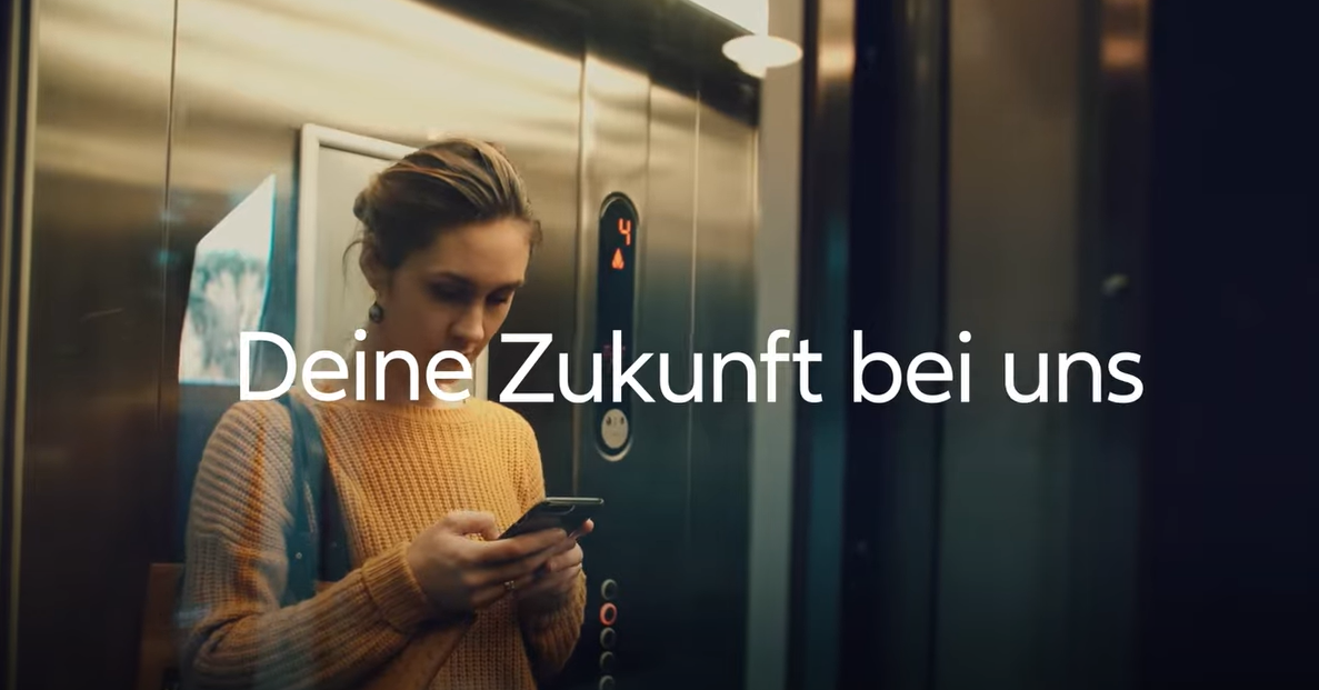 Frau steht mit ihrem Handy im Aufzug und trägt einen gelben Pullover. Neben ihr steht der Schriftzug Deine Zukunft bei uns.