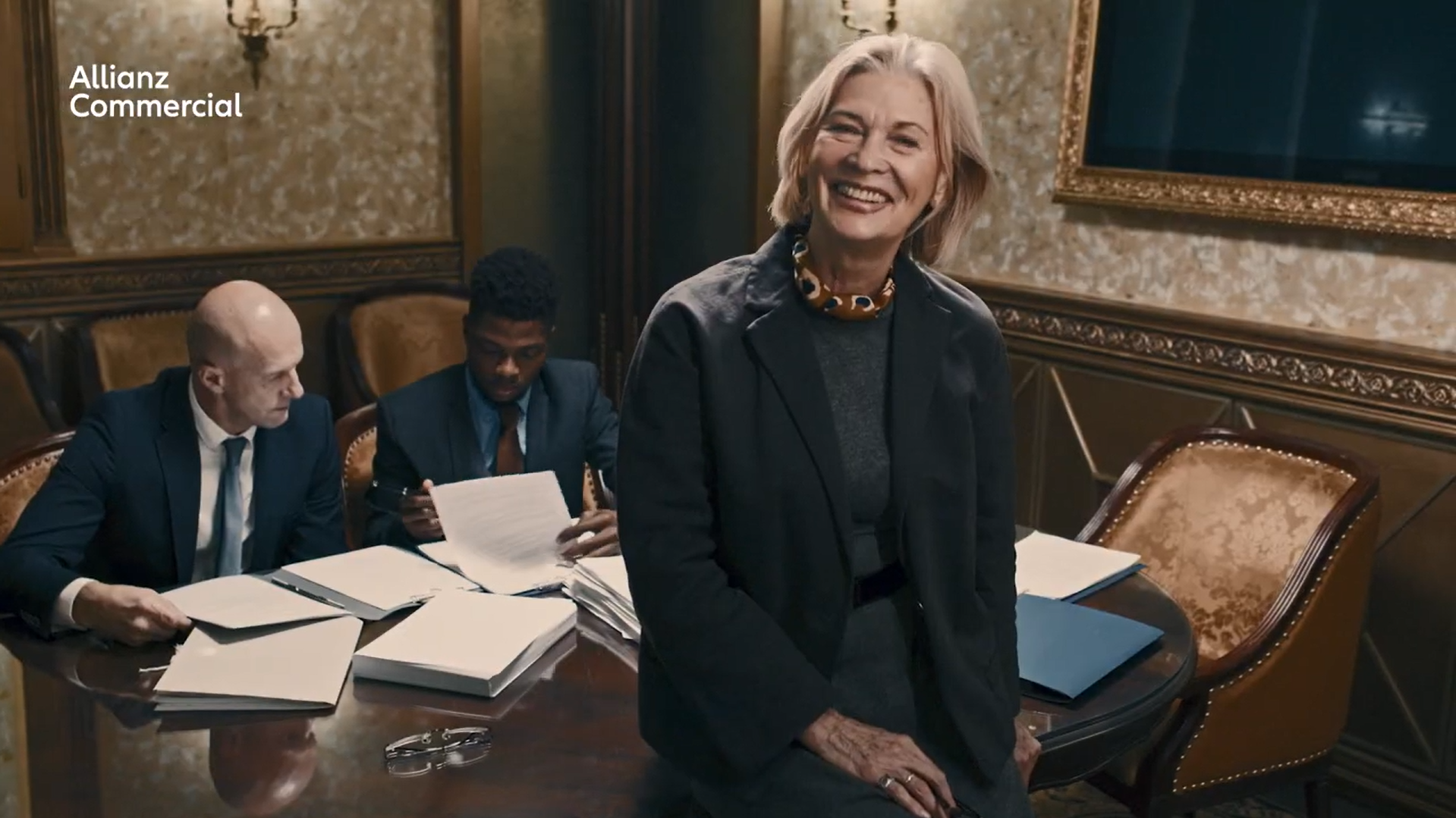 A happy woman smiling and looking directly at the camera, with her colleagues in the background focused on verifying some papers