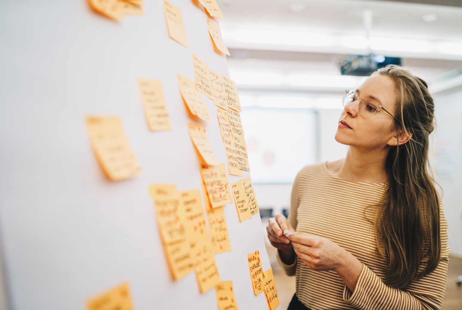 A woman wearing glasses is looking at a whiteboard with yellow sticky notes