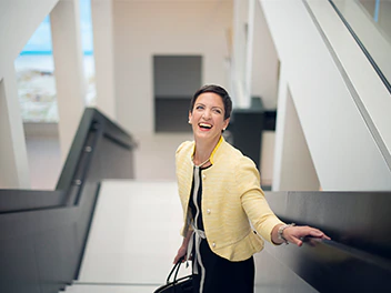 A woman in a yellow blazer smiles while looking back as she rides down the escalator