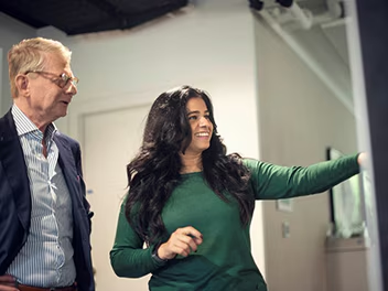 A woman in a green sweater points at a screen while a man in a suit and glasses observes. They are in a modern office environment, and both appear engaged in their discussion.