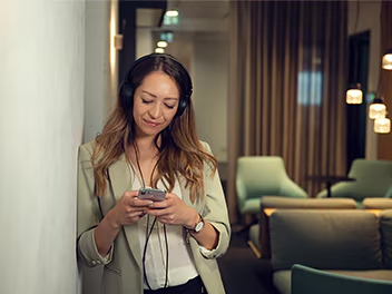 A woman in a blazer and headphones leans against a wall, looking at her phone, in a modern lounge area with soft seating and pendant lights
