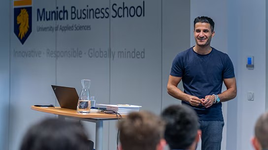 Speaker in a blue t-shirt presenting in a classroom, with 'Munich Business School' branding visible in the background