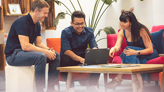 Colleagues collaborating with each other in a modern workplace, gathered around a desk, discussing documents and using a laptop.