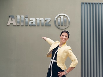 A cheerful Allianz employee stands in front of a building displaying the Allianz logo prominently.