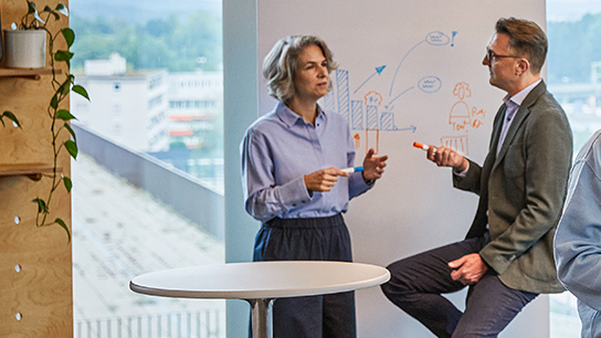 Two experienced colleagues sit opposite each other in a meeting room and talk.
