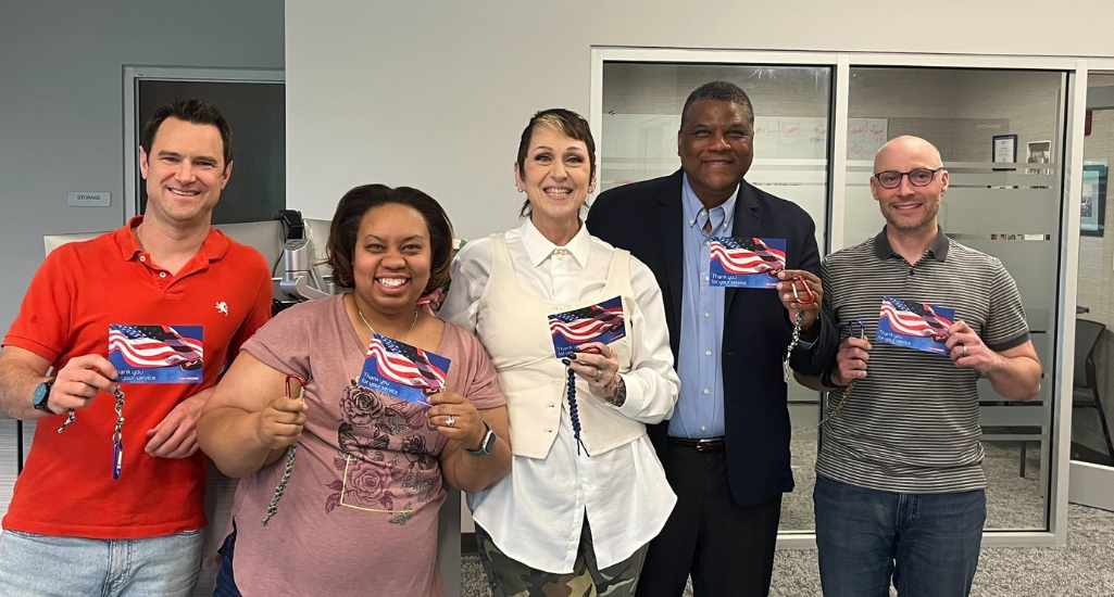 Five employees stand together in an office, smiling and holding “Thank you for your service” cards and braided lanyards during a community investment recognition moment honoring veterans and service members.