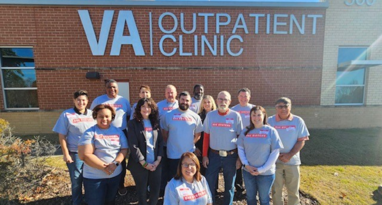 A group of 13 BAE Systems volunteers wearing matching gray company T-shirts pose outside a VA Outpatient Clinic during a community investment event supporting veterans.