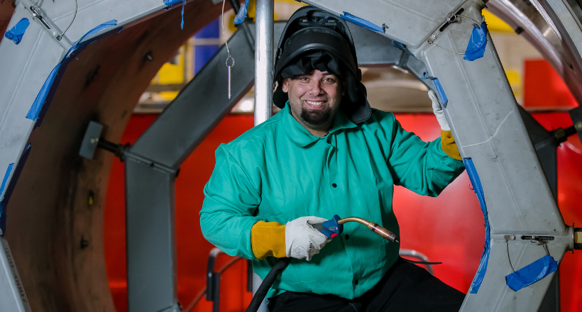A BAE Systems welder works with focused precision in an industrial setting, surrounded by heavy steel structures and large-scale equipment. Dressed in protective gear, the welder represents the craftsmanship, safety, and pride behind work that helps protect those who serve.