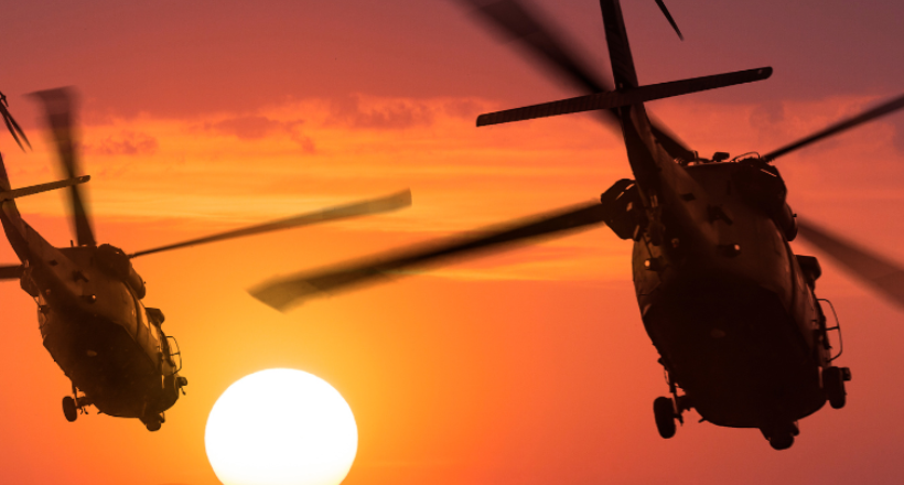Two helicopters fly into a sunset.