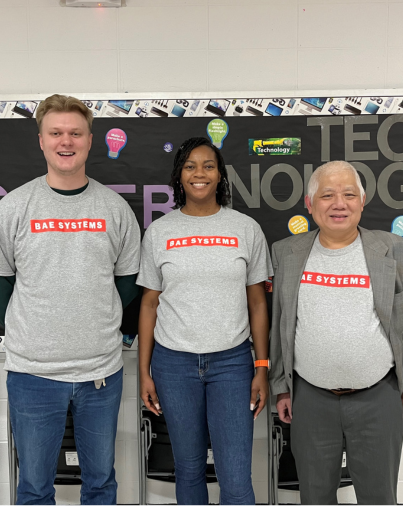 Three BAE Systems volunteers stand in front of a classroom technology bulletin board during a community investment event, smiling and wearing matching company shirts.