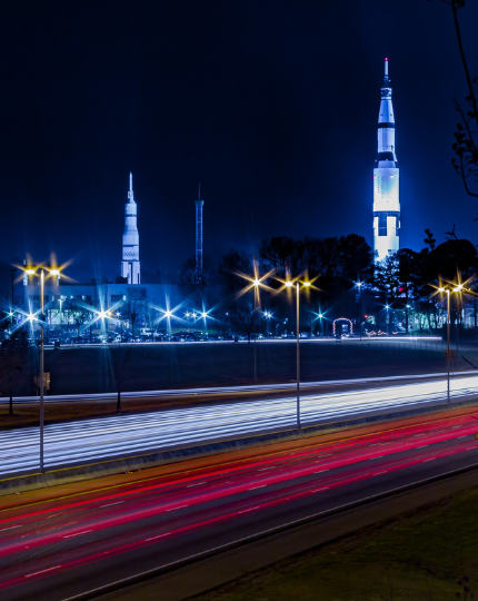 Long-exposure night view of traffic passing the U.S. Space & Rocket Center in Huntsville, with illuminated rockets rising above the highway.