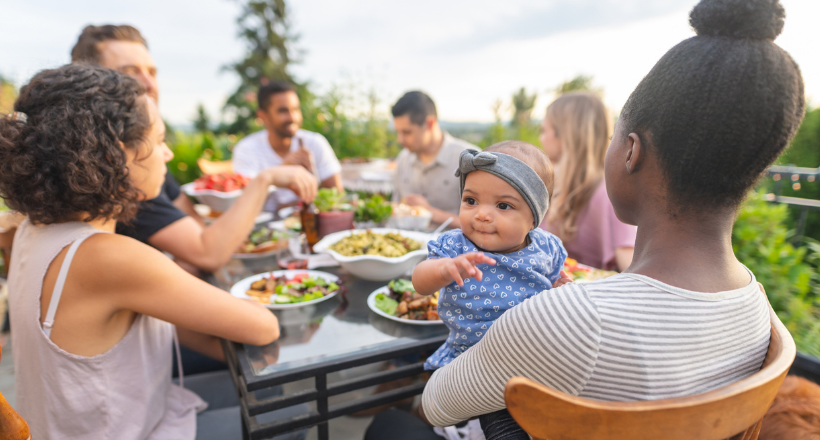 A multiethnic group of young friends enjoy good food and conversation together on a terrace outside on a summer evening. The focus is on an adorable young girl who's smiling while mom holds her.