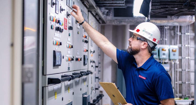 A BAE Systems program manager wearing a white hard hat, safety glasses, and a navy polo shirt adjusts a large industrial control panel while holding a clipboard in a mechanical room. The image highlights hands-on technical expertise, safety, and precision, with switches, gauges, and indicator lights reinforcing a professional industrial environment.